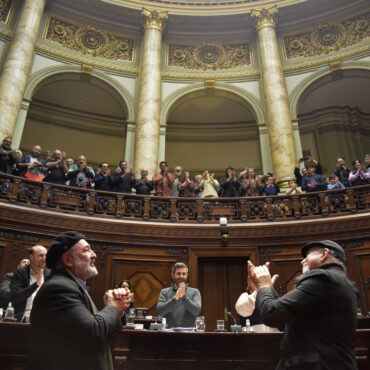 Eduardo Larbanois y Mario Carrero de pie en la Cámara de Senadores aplaudiendo observan a los senadores y a la barra que los aplauden también.
