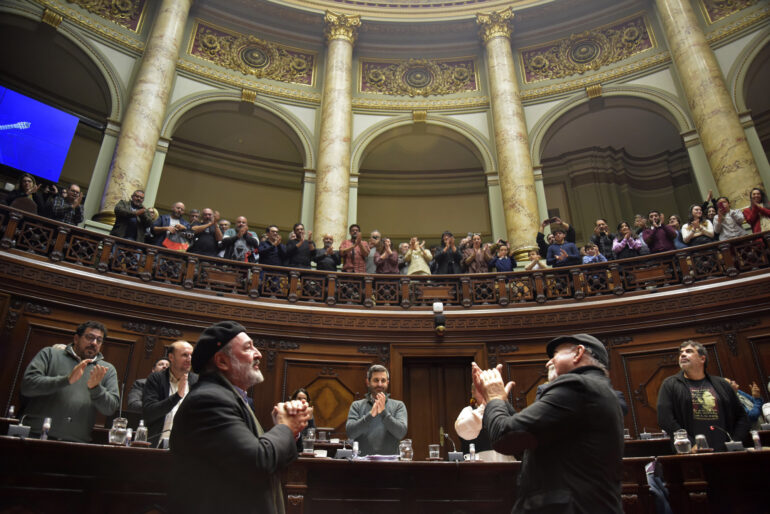 Eduardo Larbanois y Mario Carrero de pie en la Cámara de Senadores aplaudiendo observan a los senadores y a la barra que los aplauden también.
